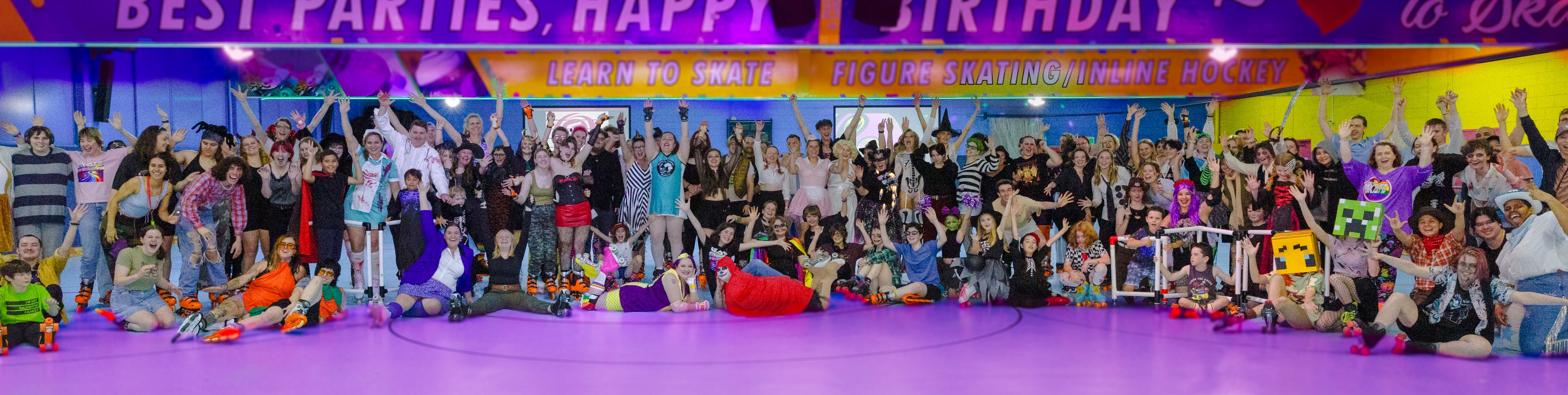 A large, colorful group of people gathers on a vibrant roller skating rink for a joyful celebration. Some are wearing skates while others stand or sit, striking cheerful poses. Banners overhead advertise parties, skating, and hockey lessons, adding to the festive atmosphere.