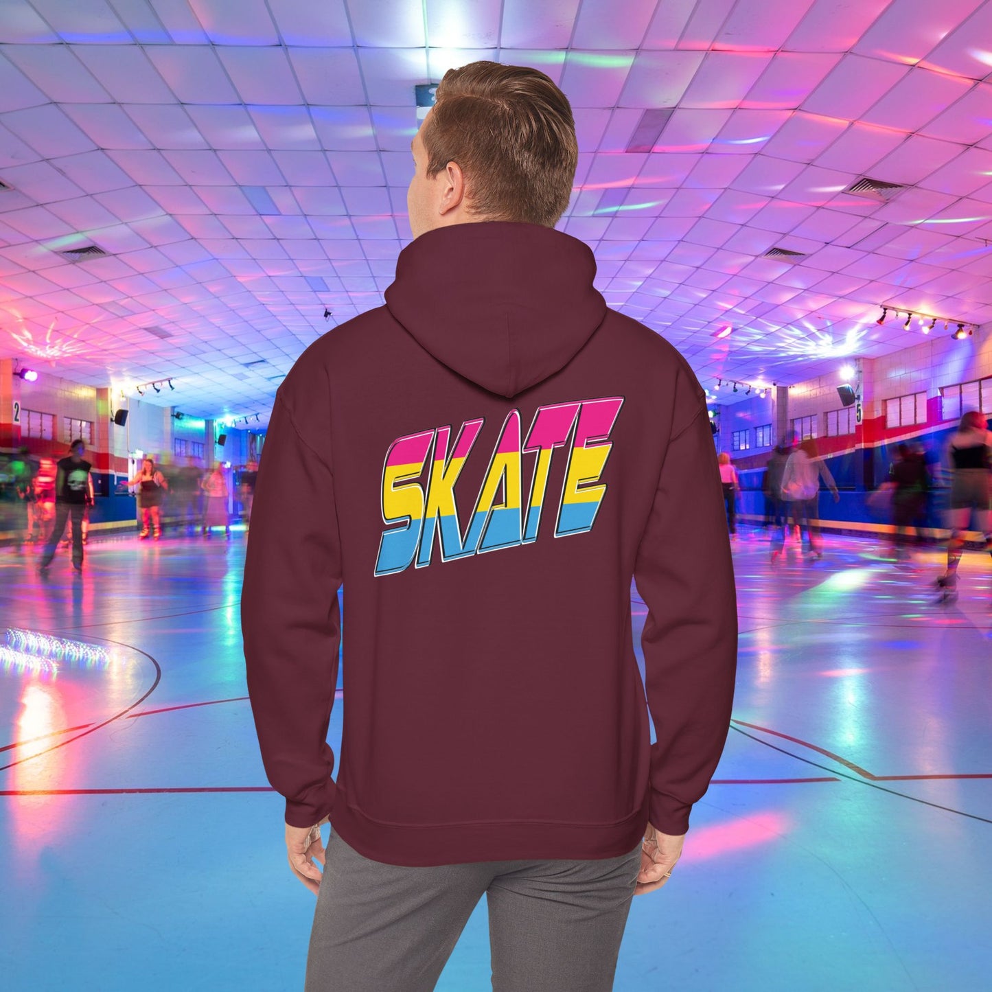 A person wearing a high-quality SKATE Pansexual Pride Hoodie by Printify, featuring the word "SKATE" in large, colorful letters, stands in a vibrant roller skating rink. The unisex heavy blend hoodie is part of a trans and queer-owned brand. The rink is decorated with colorful lights, and people are skating in the background as the person faces away from the camera.