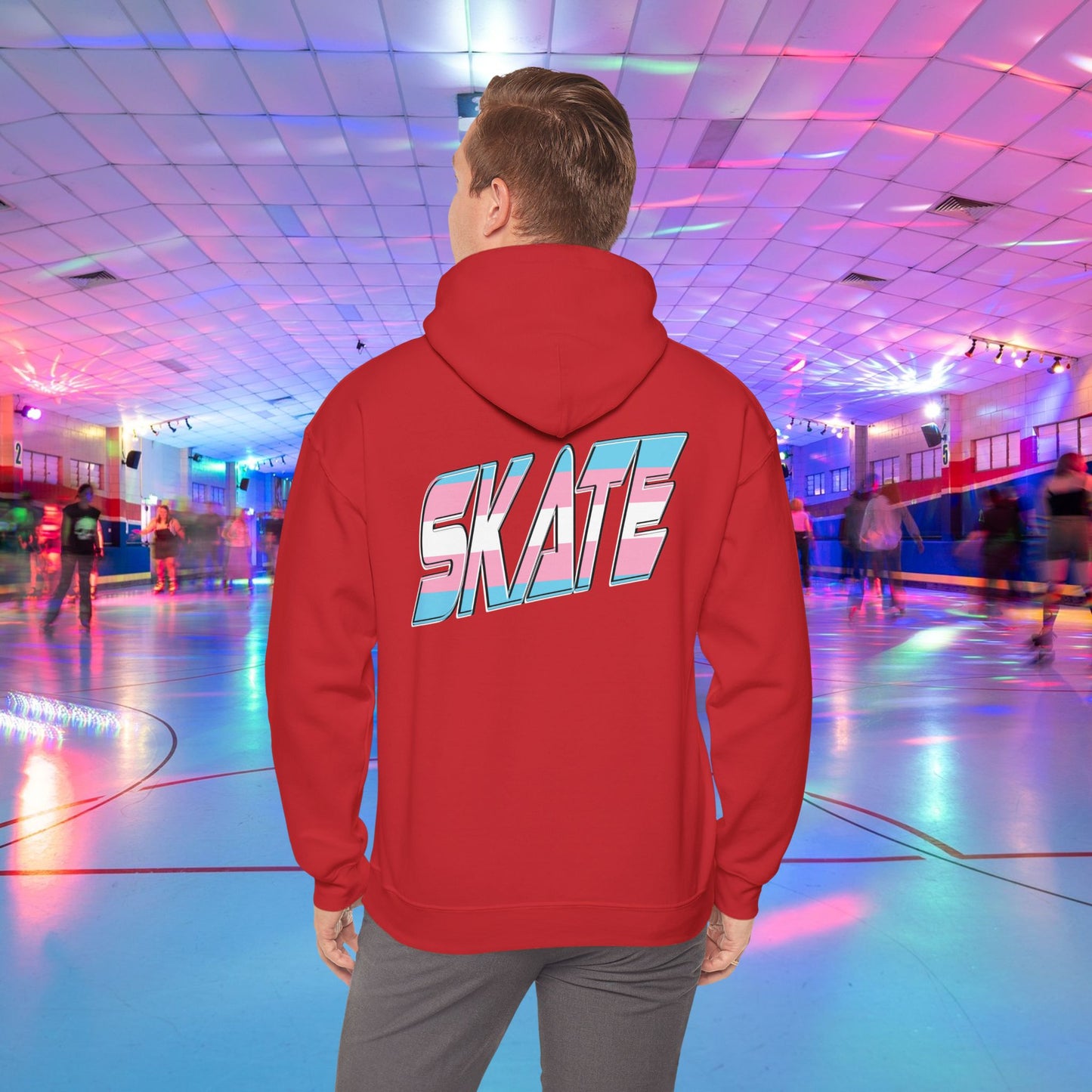 A man with light brown hair, wearing a black hoodie that features "SKATE" in large, colorful letters on the back, stands in a roller skating rink illuminated by vibrant lighting with people skating in the background. The high-quality SKATE Trans Flag Hoodie is made from ethically grown US cotton and is part of Printify's unisex heavy blend collection. Available for Australian shipping.