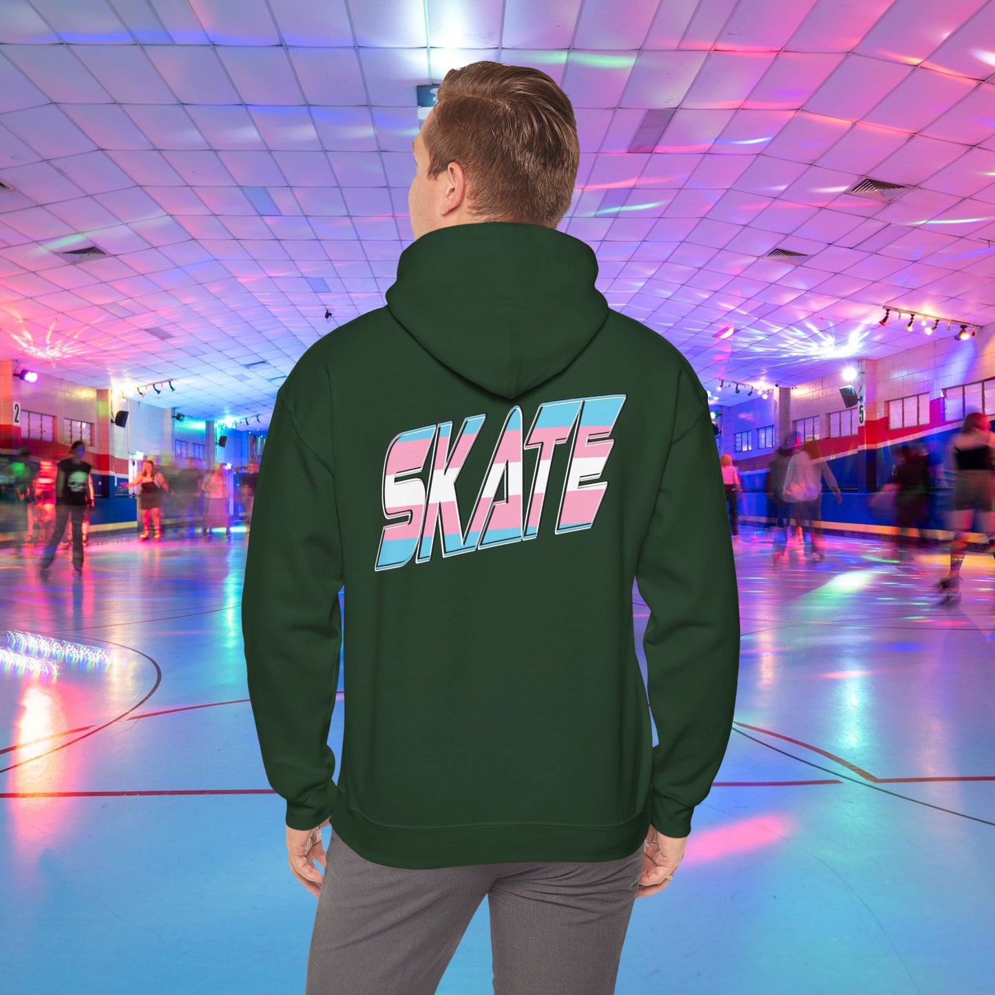 A man with light brown hair, wearing a black hoodie that features "SKATE" in large, colorful letters on the back, stands in a roller skating rink illuminated by vibrant lighting with people skating in the background. The high-quality SKATE Trans Flag Hoodie is made from ethically grown US cotton and is part of Printify's unisex heavy blend collection. Available for Australian shipping.