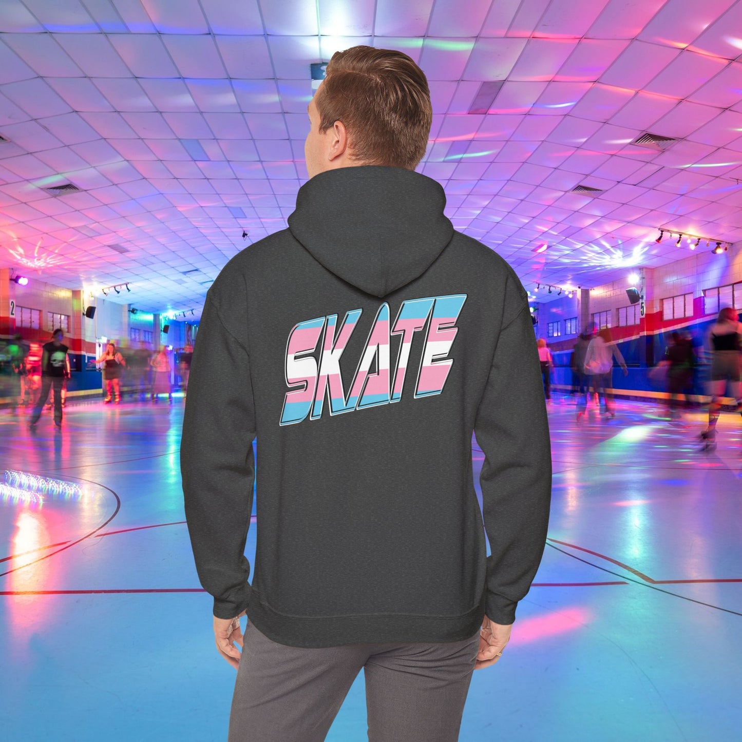 A person wearing a SKATE Transgender Pride Hoodie by Printify, featuring bold, colorful letters on the back, stands in a brightly lit roller skating rink. The background showcases people skating under vibrant, multicolored lights, creating a lively atmosphere.