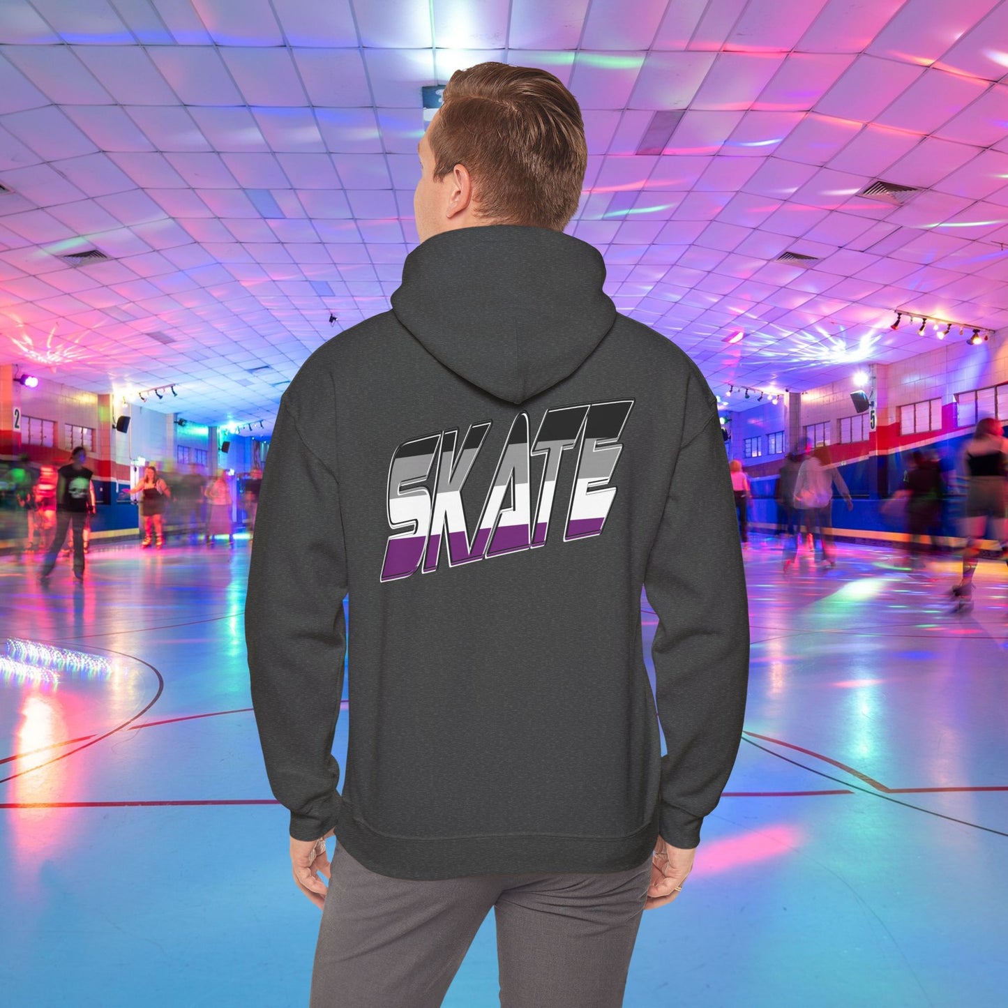 A person wearing a Printify SKATE Asexual Pride Hoodie, featuring the word "SKATE" printed on the back in gradient colors, stands in a brightly lit roller skating rink, observing the skaters. The rink is illuminated with colorful lights and exudes a lively atmosphere.