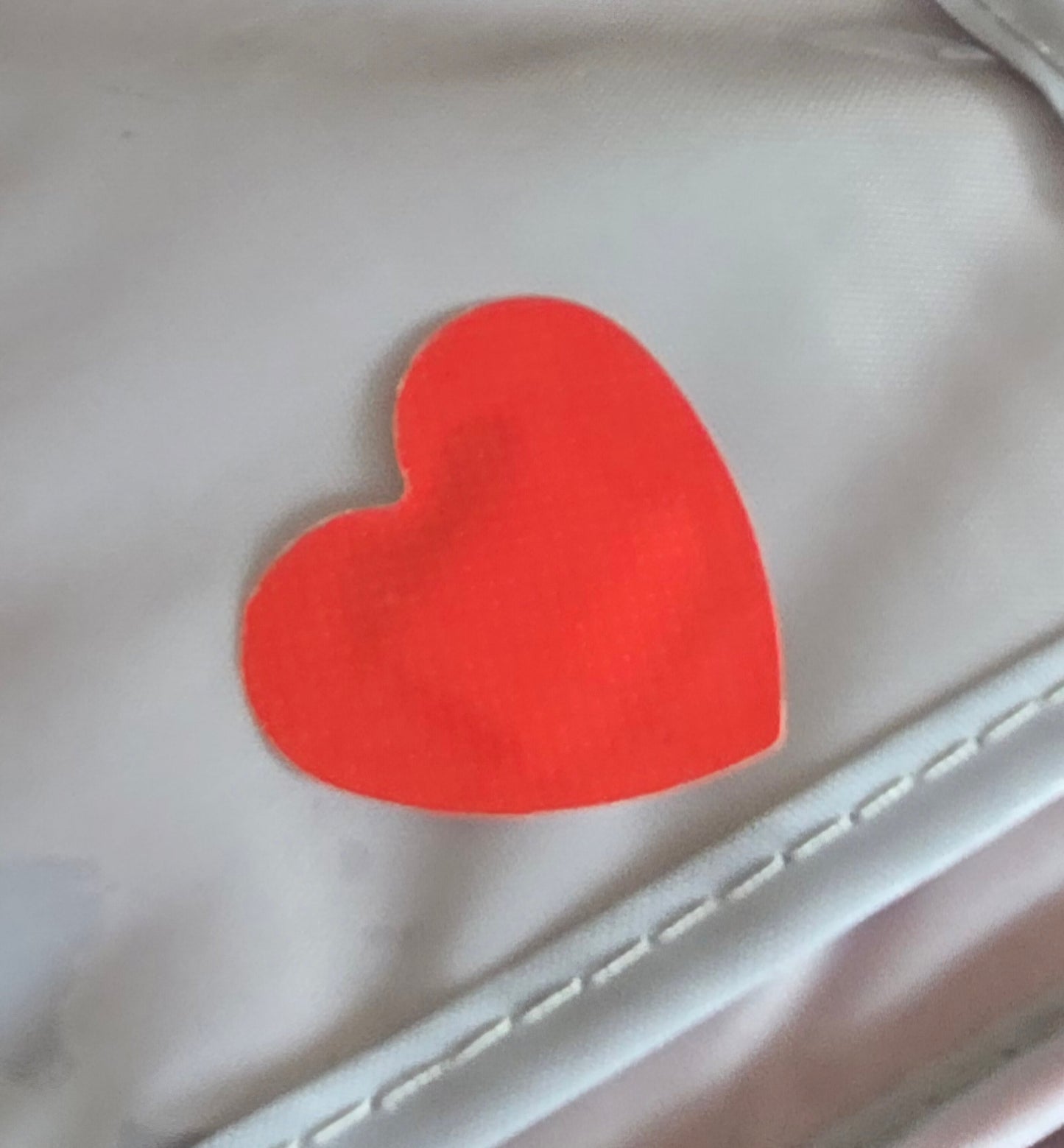 A red, heart-shaped band-aid from the "20 love-heart shaped red Band Aids" set is on a light gray surface with visible stitching along the edges, resembling graze dressings. Its texture contrasts with the smooth background.