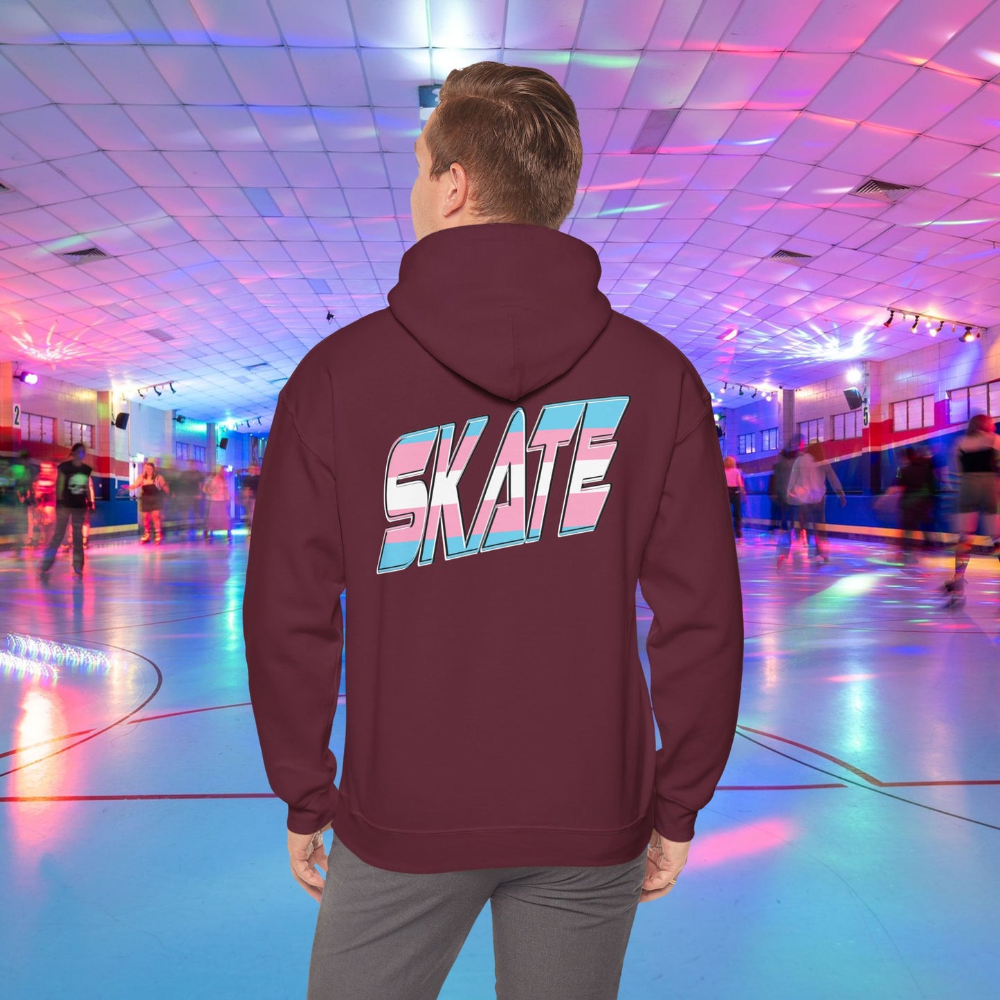 A person wearing a SKATE Transgender Pride Hoodie by Printify, featuring bold, colorful letters on the back, stands in a brightly lit roller skating rink. The background showcases people skating under vibrant, multicolored lights, creating a lively atmosphere.