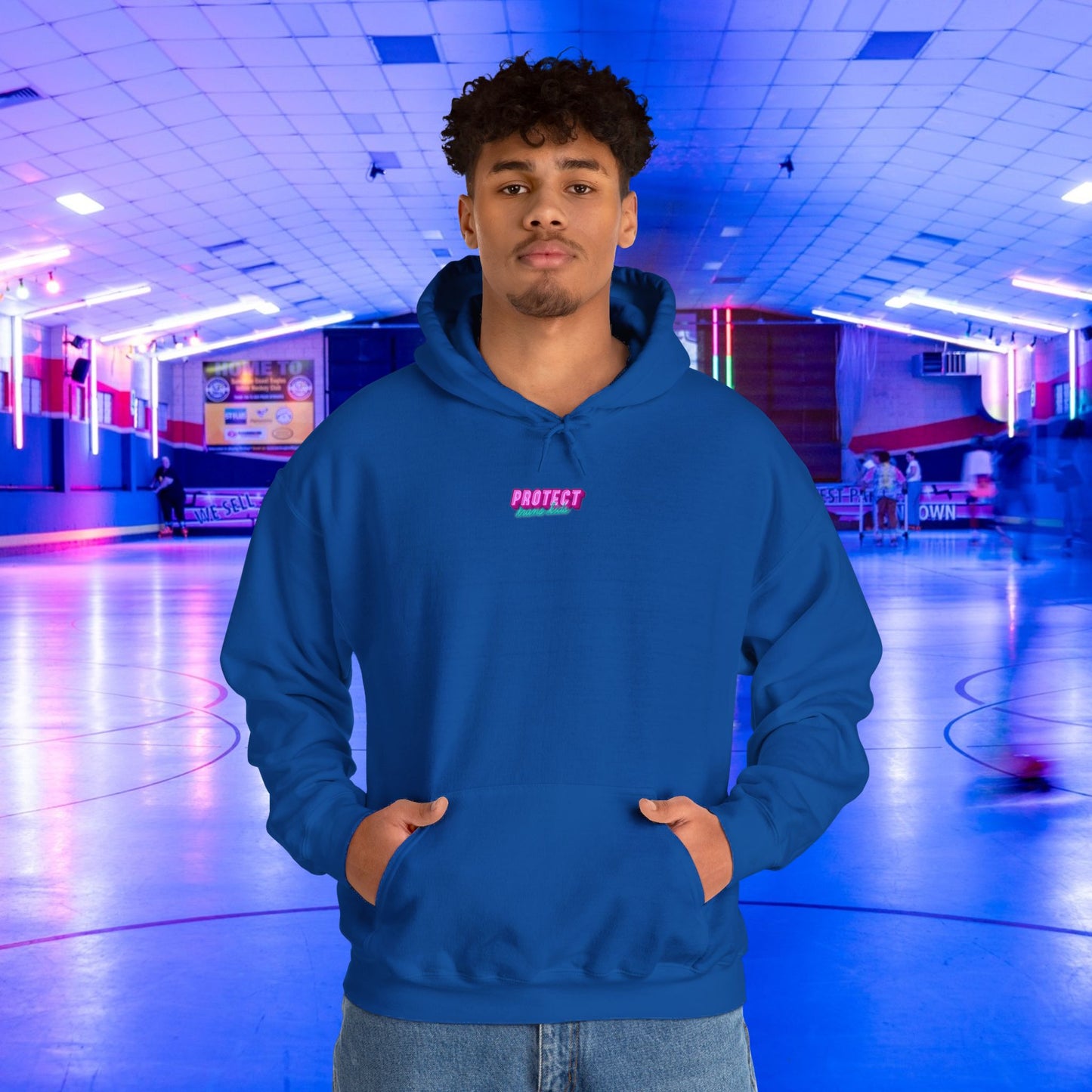 Standing confidently in a vibrant, neon-lit indoor skating rink, a person dons the Protect Trans Kids Hoodie - Australian Shipping. The ethically grown cotton unisex sweatshirt features "PROTECT" in pink across the chest, adding flair to the lively atmosphere.