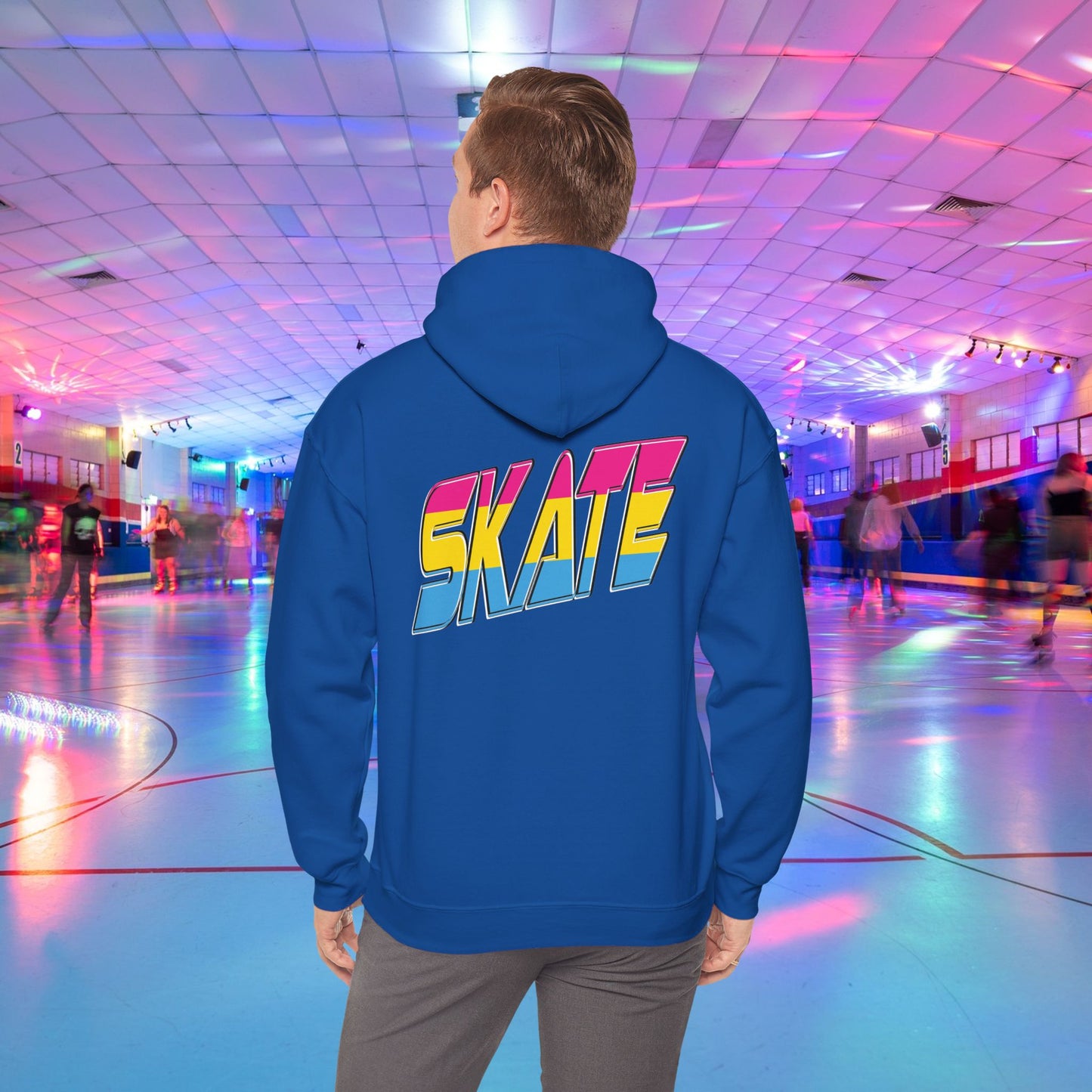 A person wearing a high-quality SKATE Pansexual Pride Hoodie by Printify, featuring the word "SKATE" in large, colorful letters, stands in a vibrant roller skating rink. The unisex heavy blend hoodie is part of a trans and queer-owned brand. The rink is decorated with colorful lights, and people are skating in the background as the person faces away from the camera.