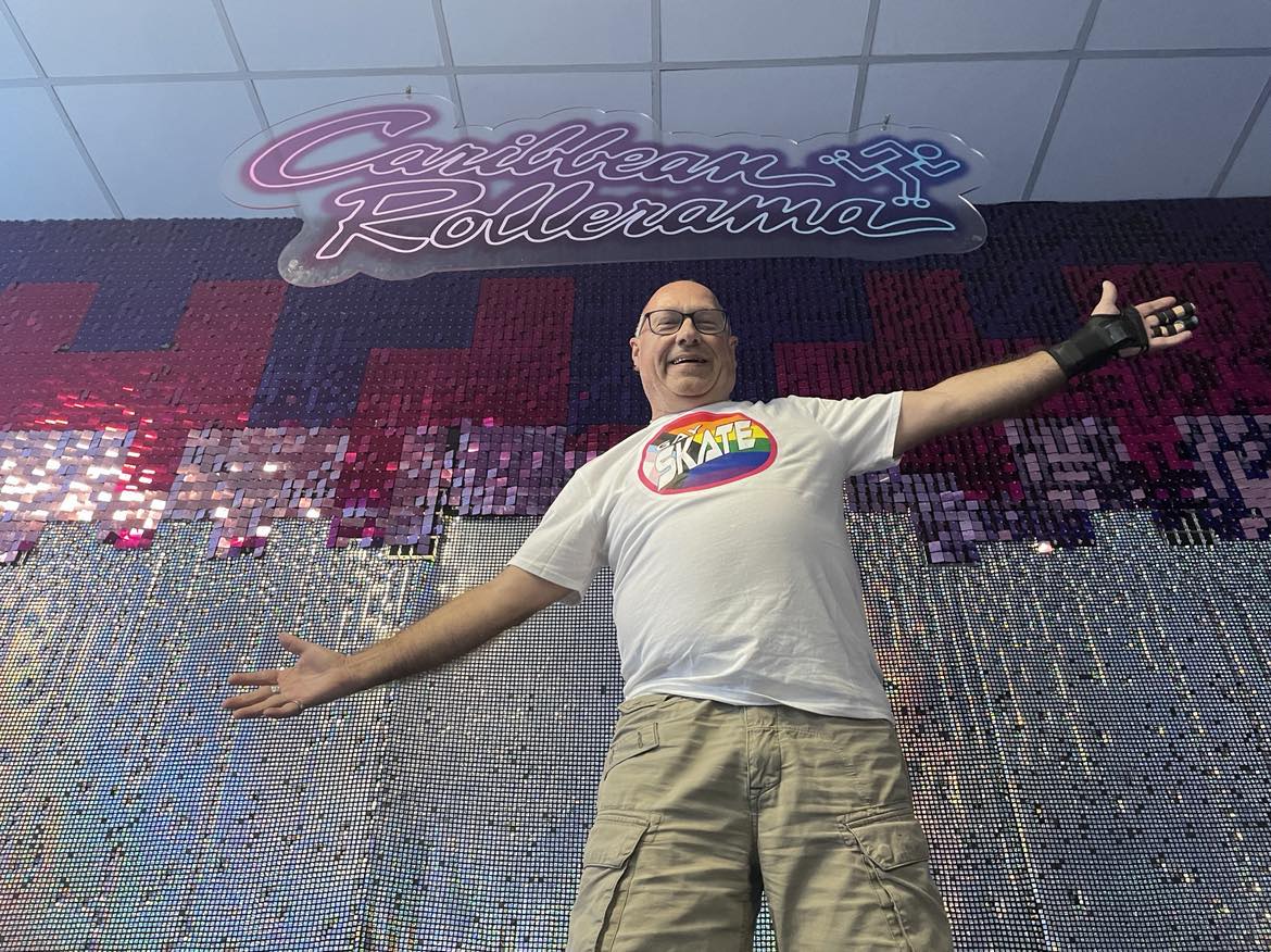A smiling man stands with arms outstretched in front of a sparkly pink and purple backdrop with a sign that reads "Caribbean Rollerrama." He is wearing glasses, a white T-shirt with a rainbow-colored "SKATE" logo, beige shorts, and a wrist guard.