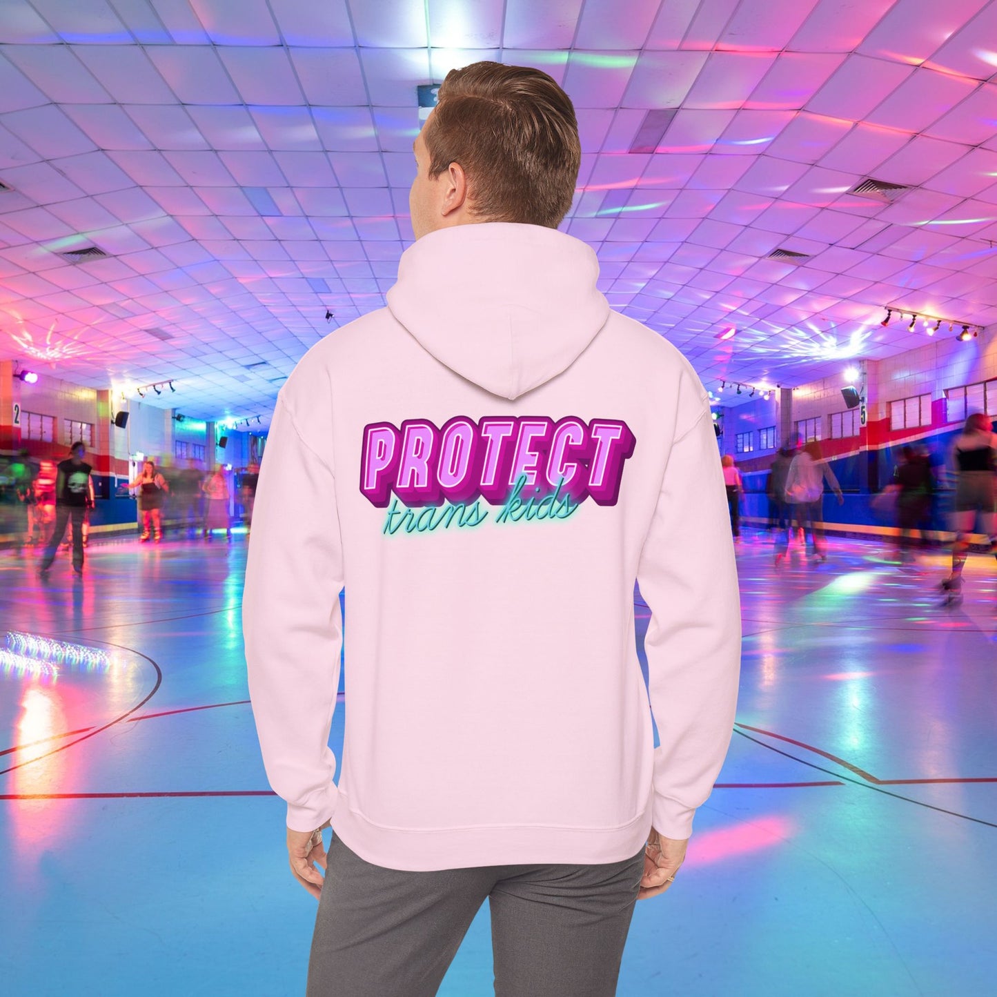 A person is at a roller skating rink, sporting a high-quality Protect Trans Kids Hoodie from Printify, crafted from ethically grown cotton, with "PROTECT trans kids" boldly and colorfully emblazoned on the back. The rink is bathed in vibrant, colorful lights, and other skaters can be seen gliding in the background.
