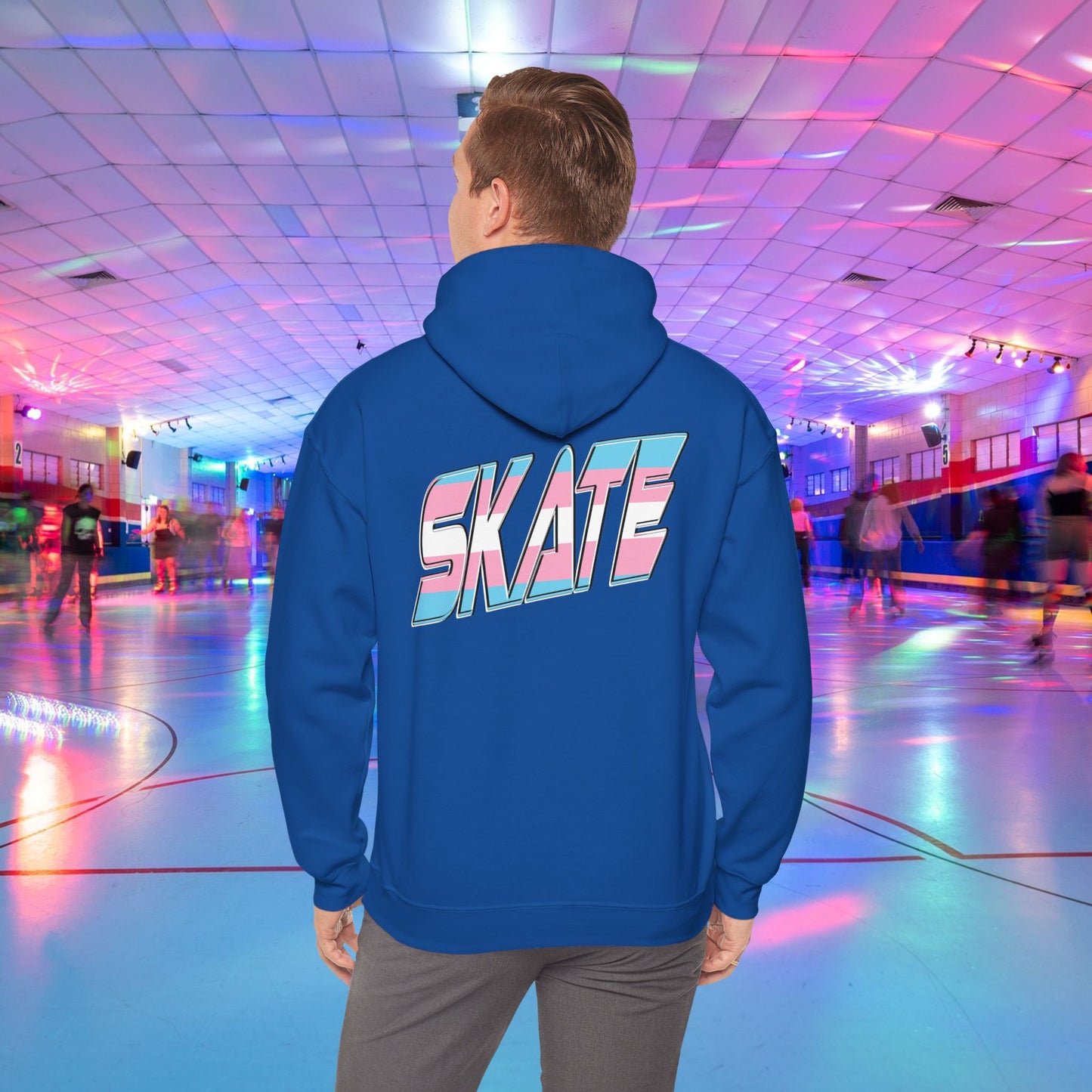 A man with light brown hair, wearing a black hoodie that features "SKATE" in large, colorful letters on the back, stands in a roller skating rink illuminated by vibrant lighting with people skating in the background. The high-quality SKATE Trans Flag Hoodie is made from ethically grown US cotton and is part of Printify's unisex heavy blend collection. Available for Australian shipping.