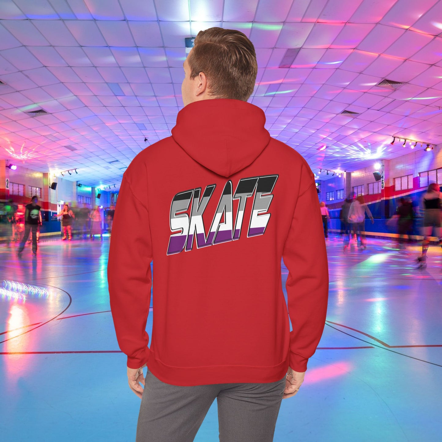 A person wearing a Printify SKATE Asexual Pride Hoodie, featuring the word "SKATE" printed on the back in gradient colors, stands in a brightly lit roller skating rink, observing the skaters. The rink is illuminated with colorful lights and exudes a lively atmosphere.