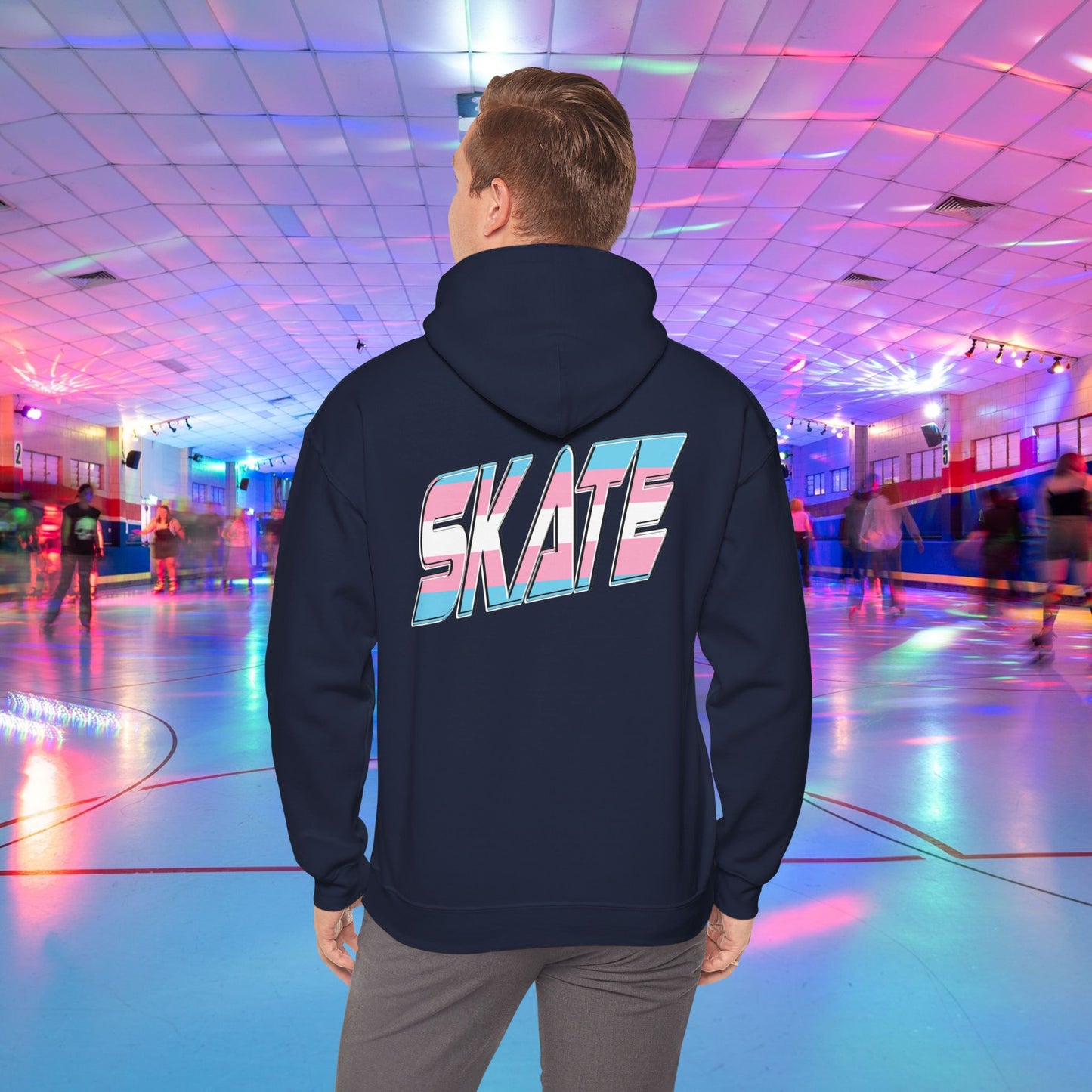 A person wearing a SKATE Transgender Pride Hoodie by Printify, featuring bold, colorful letters on the back, stands in a brightly lit roller skating rink. The background showcases people skating under vibrant, multicolored lights, creating a lively atmosphere.