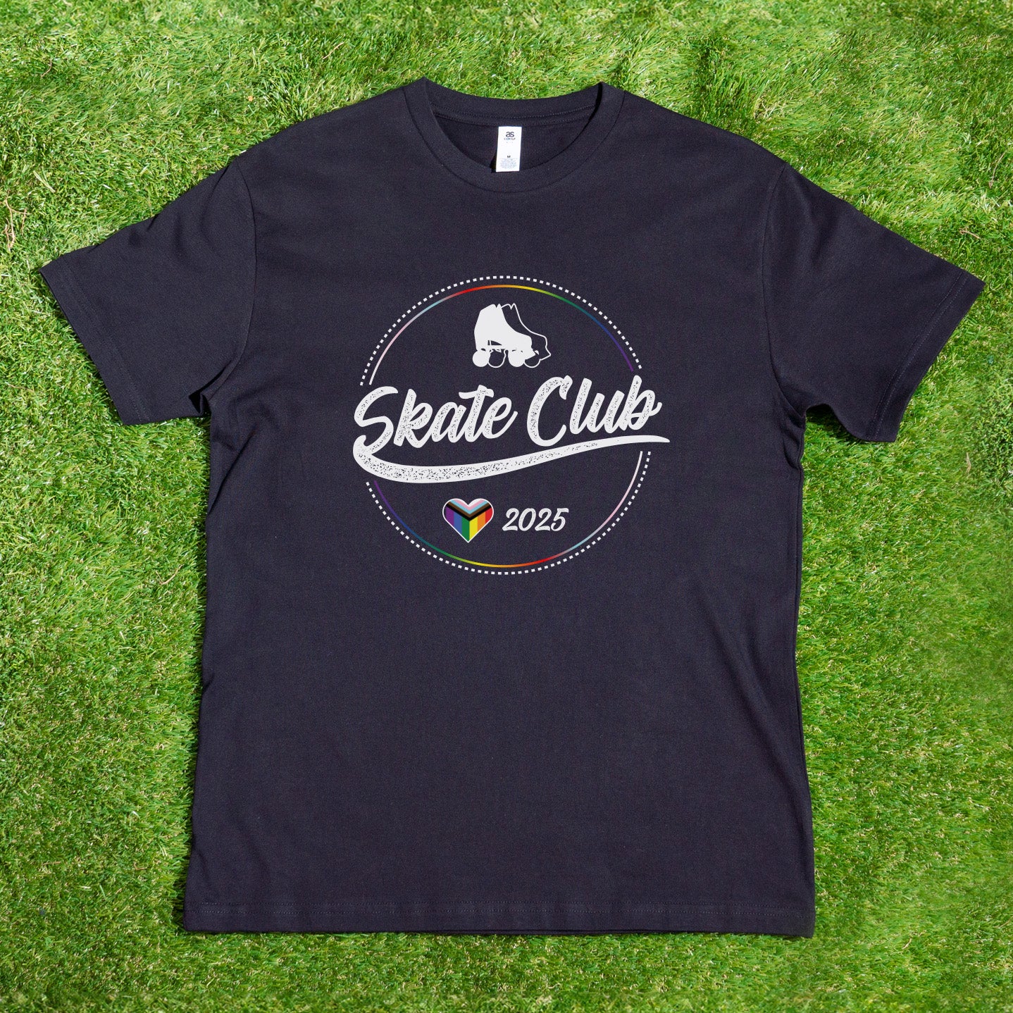 Laid flat on green grass, the Gay Skate "Pre-order: SKATE CLUB 2025 Progress Pride" T-shirt features a circular logo with a roller skate and "Skate Club 2025" text. Below, a rainbow-colored heart symbolizes inclusion, celebrating its roots as a queer and trans-owned business.