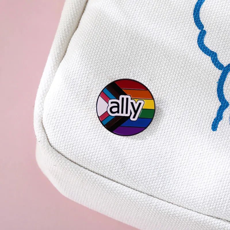 The Ally enamel pin featuring progress pride flag, with a rainbow design and “ally” in white text, is shown attached to a white bag with blue stitching against a pink background—an uplifting addition to your pride collection.