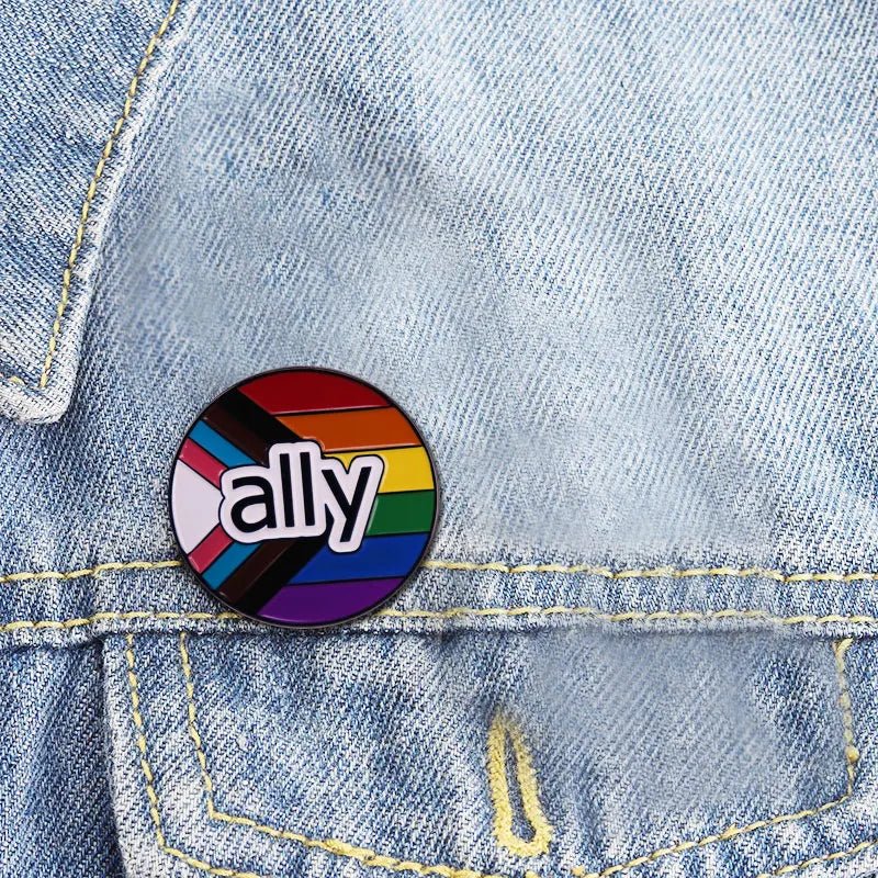 The Ally enamel pin featuring the Progress Pride Flag displays "ally" atop intersecting black, brown, pink, blue, white, and rainbow stripes. A bold pride accessory shown on light blue denim fabric.