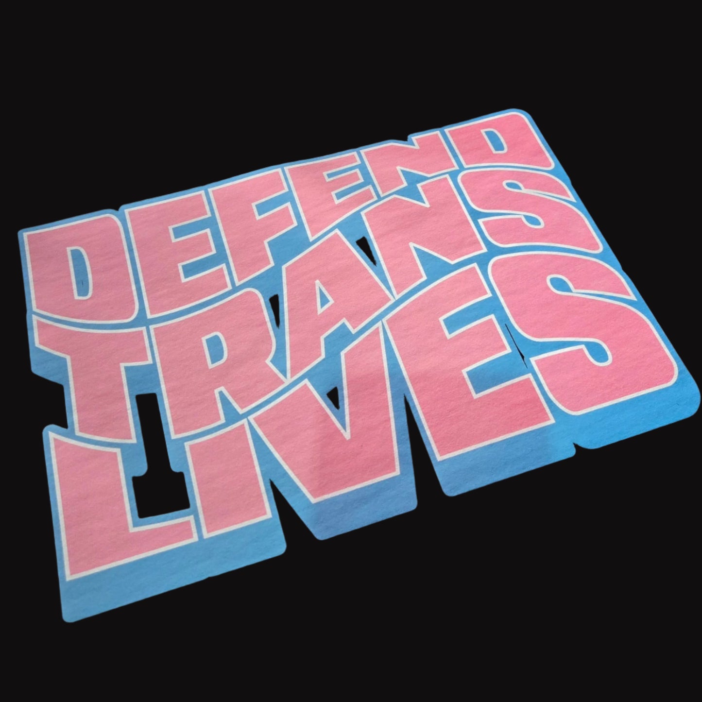 The "Defend Trans Lives - Local hand printed t-shirt" features bold blue and pink text on black, inspired by the trans pride flag. Each shirt is hand printed locally, making every piece unique.