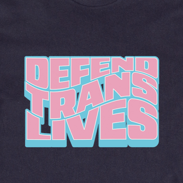 A "Defend Trans Lives" black T-shirt, hand printed locally with bold pink and blue letters, rests on lush green grass, embracing diversity.