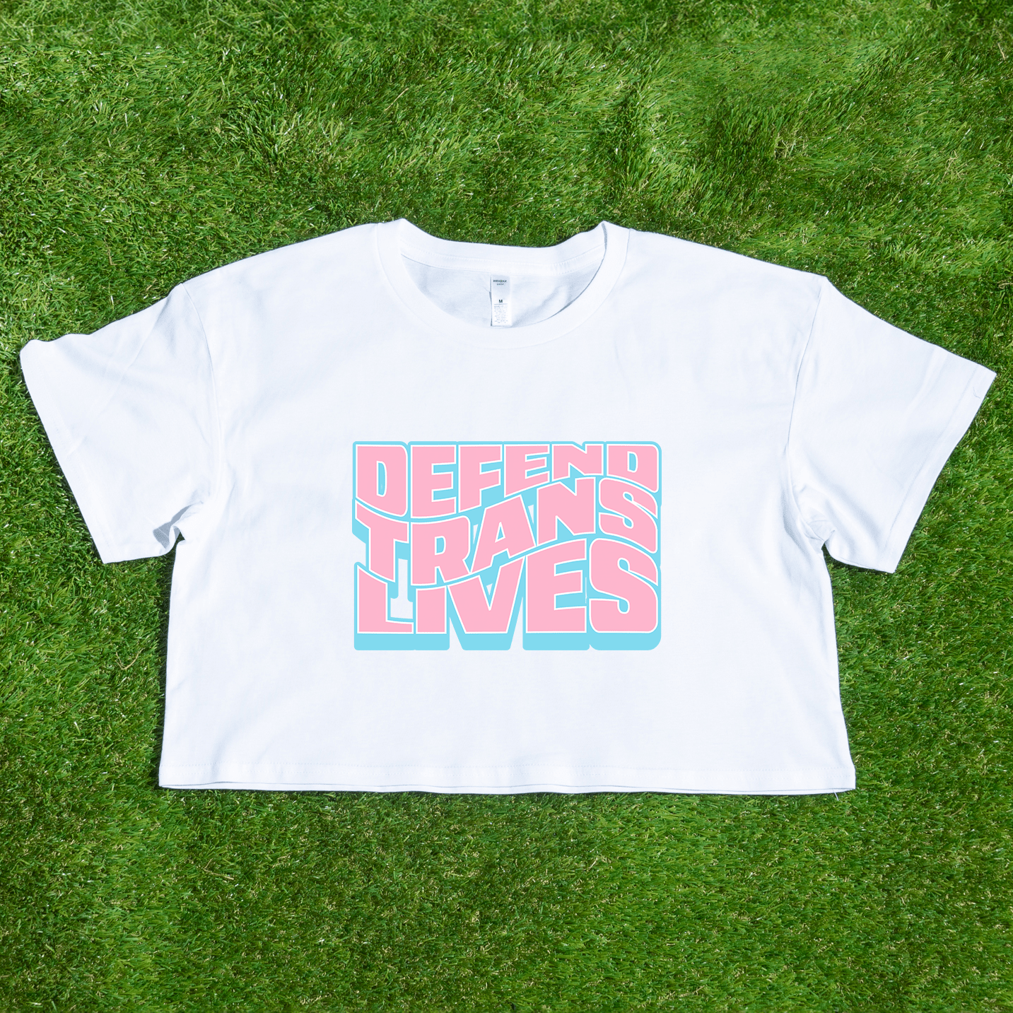 White crop top on grass showcasing "Defend Trans Lives" in bold pink and blue, a signature queer streetwear piece from a trans-owned brand.