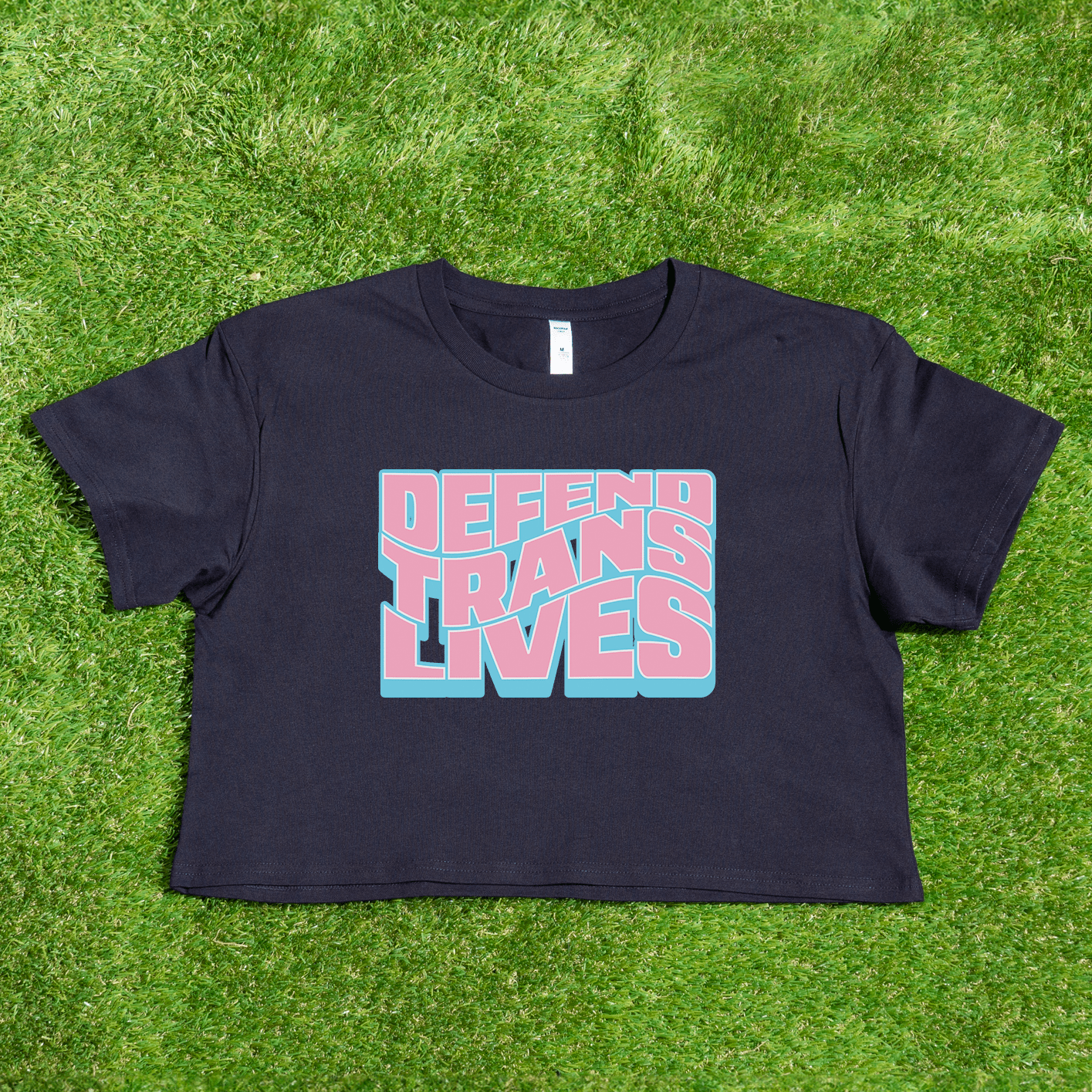 A black crop tee with "DEFEND TRANS LIVES" in vibrant pink and blue rests on the grass. This queer streetwear, named "Defend Trans Lives - original design," supports transgender pride and is locally printed by a trans-owned business.