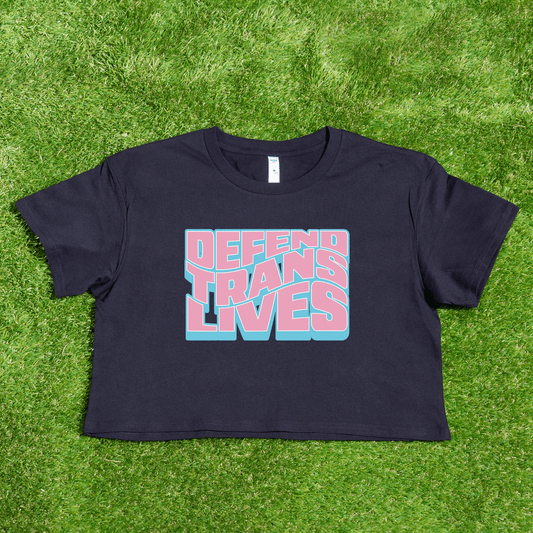 A black crop tee with "DEFEND TRANS LIVES" in vibrant pink and blue rests on the grass. This queer streetwear, named "Defend Trans Lives - original design," supports transgender pride and is locally printed by a trans-owned business.