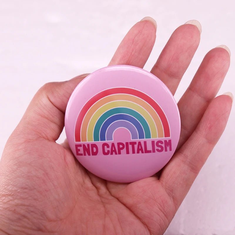 A hand holds the "End Capitalism - Pink Rainbow Button Badge," featuring a bright rainbow and bold red text. The light, neutral background accentuates this pride item's impactful message.