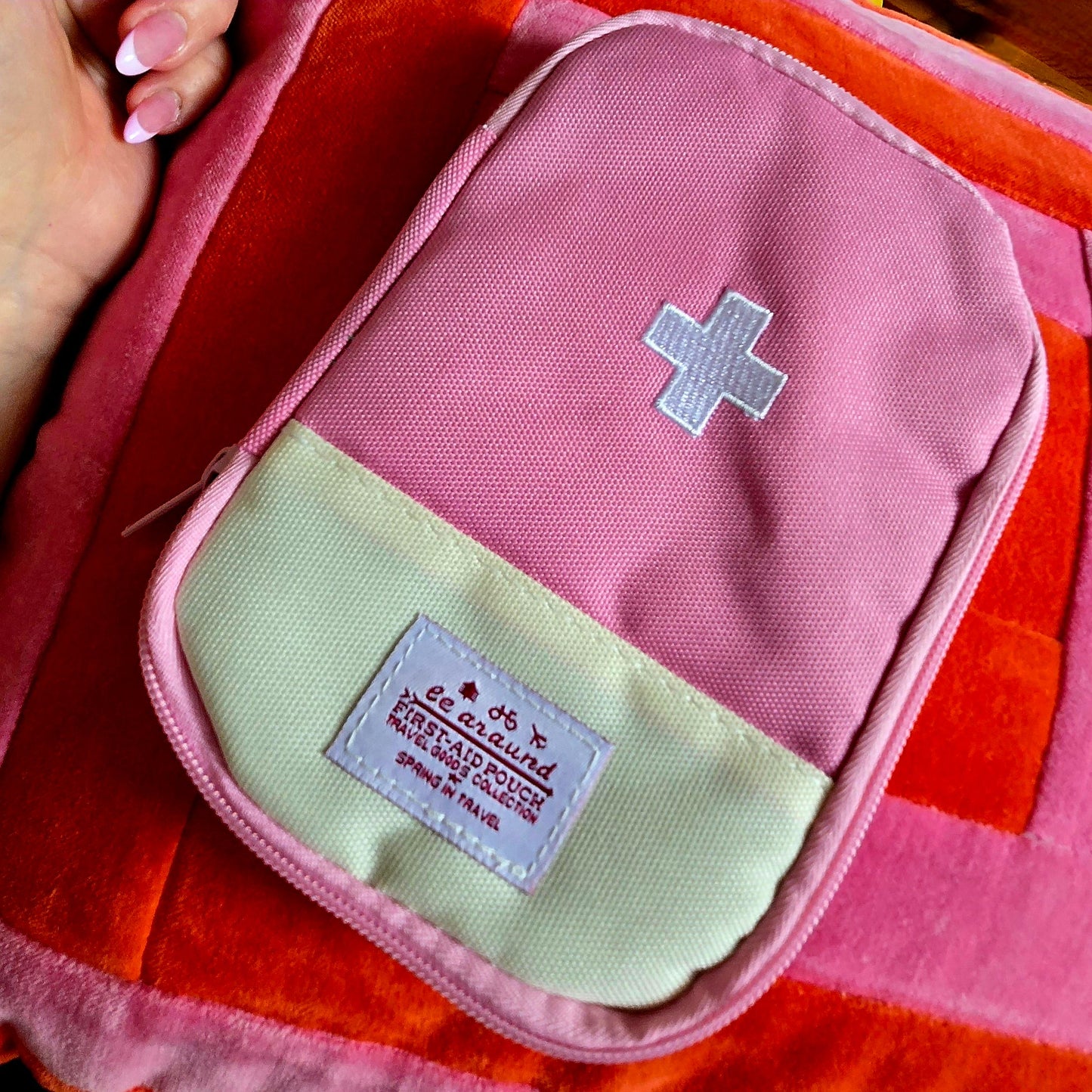 A stylish close-up features the "First Aid Kit for skate bag - cute colours and designs - case only" in pink and cream with a white cross, heart symbol, and label. Held by hand on pink and orange striped fabric, it's perfect for a compact first aid kit addition.