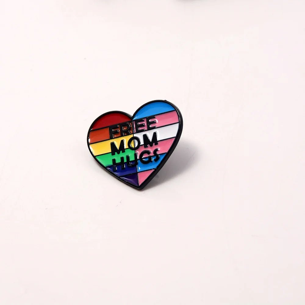A love heart badge with rainbow and trans pride flag colors and "FREE MOM HUGS" text, called the Free Mom Hugs rainbow and trans pride flag love heart badge, displayed on a plain white background.