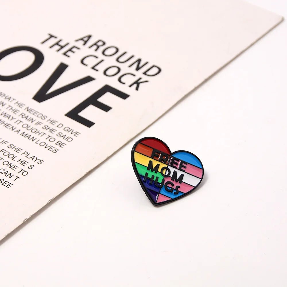 The Free Mom Hugs rainbow and trans pride flag love heart badge rests on a white surface beside a page with large black text reading "AROUND THE CLOCK LOVE.