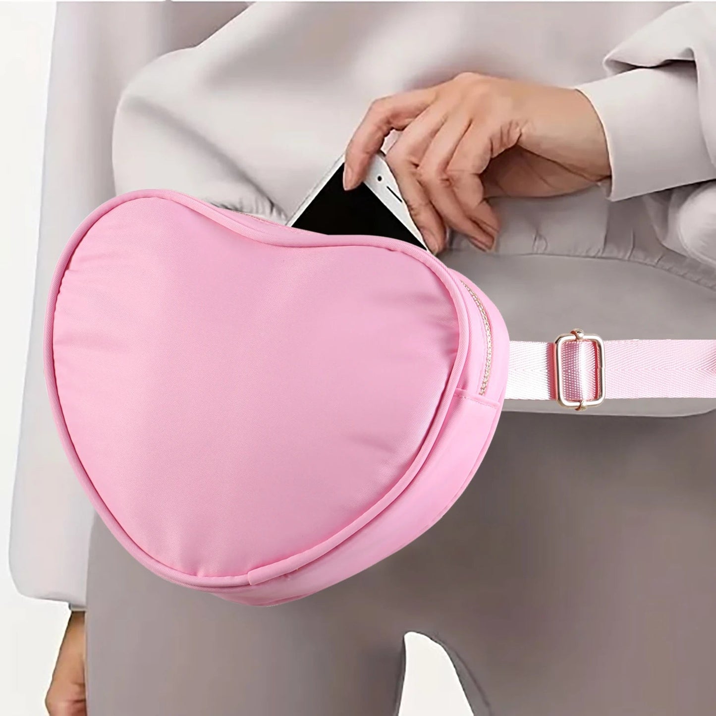 A person dressed in grey pants and a grey top adjusts a light pink, heart-shaped "Heart Shaped Crossbody Bag or Fanny Pack for skating" from Gay Skate around their waist. The individual is holding a smartphone with a white case, half inserting it into the adjustable waist strap's main compartment.