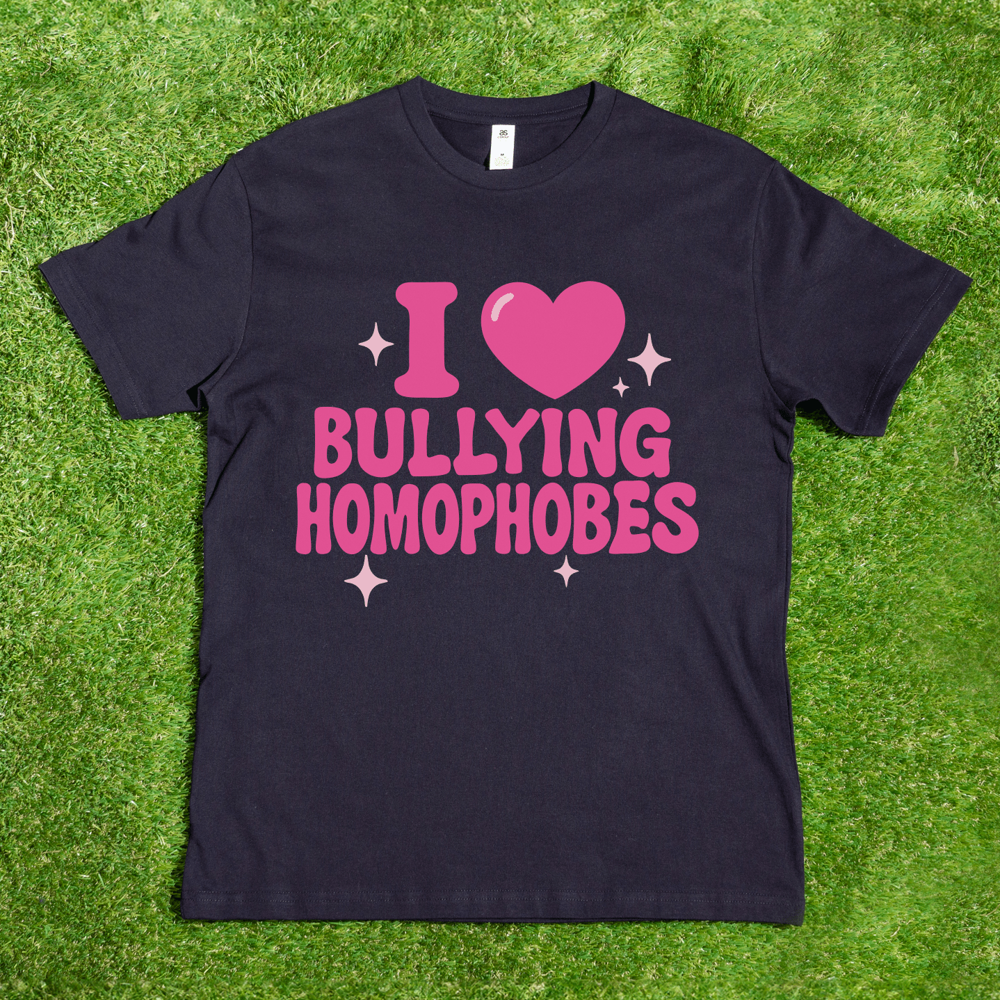 A black AS Colour tee reading "I 🩷 Bullying Transphobes / Homophobes" in pink is shown on grass, surrounded by sparkles—hand printed by a trans-owned business specializing in queer streetwear.