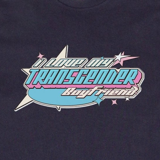 The "I LOVE MY TRANSGENDER BOYFRIEND" T-shirt from Gay Skate showcases a custom design with a vibrant retro-style graphic. The text "In Loud City, Transgender, Loud and Proud!" is prominently displayed in shades of blue, pink, and purple from the Transgender Pride Flag. The black T-shirt is laid flat on a grassy background.