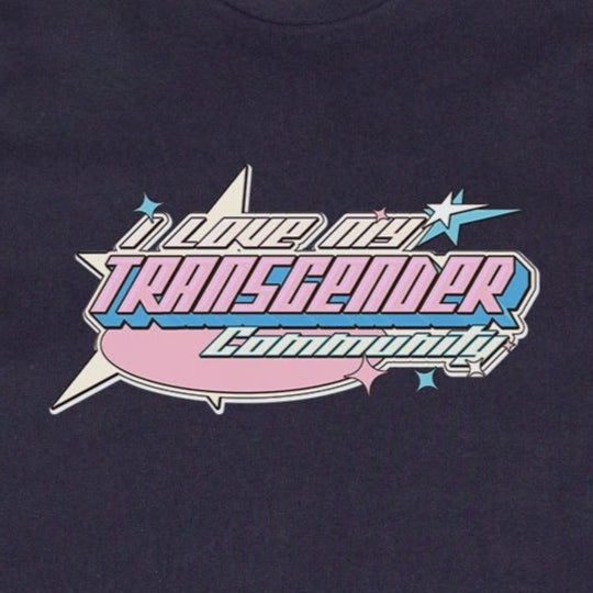 An "I LOVE MY TRANSGENDER COMMUNITY" T-shirt from Gay Skate, black in color, lying on the grass. The front boasts a vibrant pastel design with the bold, stylized text "Support Your Transgender Community" encased in a starburst shape—ideal Pride clothing from a queer and trans owned business.