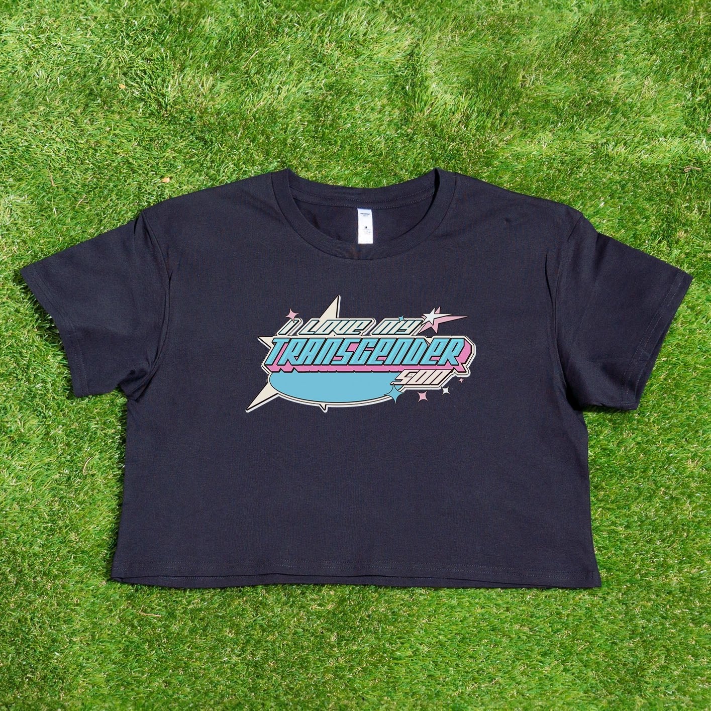 A pink crop top from Gay Skate, emblazoned with "Proud Transgender Daughter" in bold text and decorative elements, lies flat on green grass. This shirt, part of the "I LOVE MY TRANSGENDER DAUGHTER or SON" collection, boasts a classic short-sleeve design and is one of our unique original designs from a queer-owned business. Available for Australian shipping.