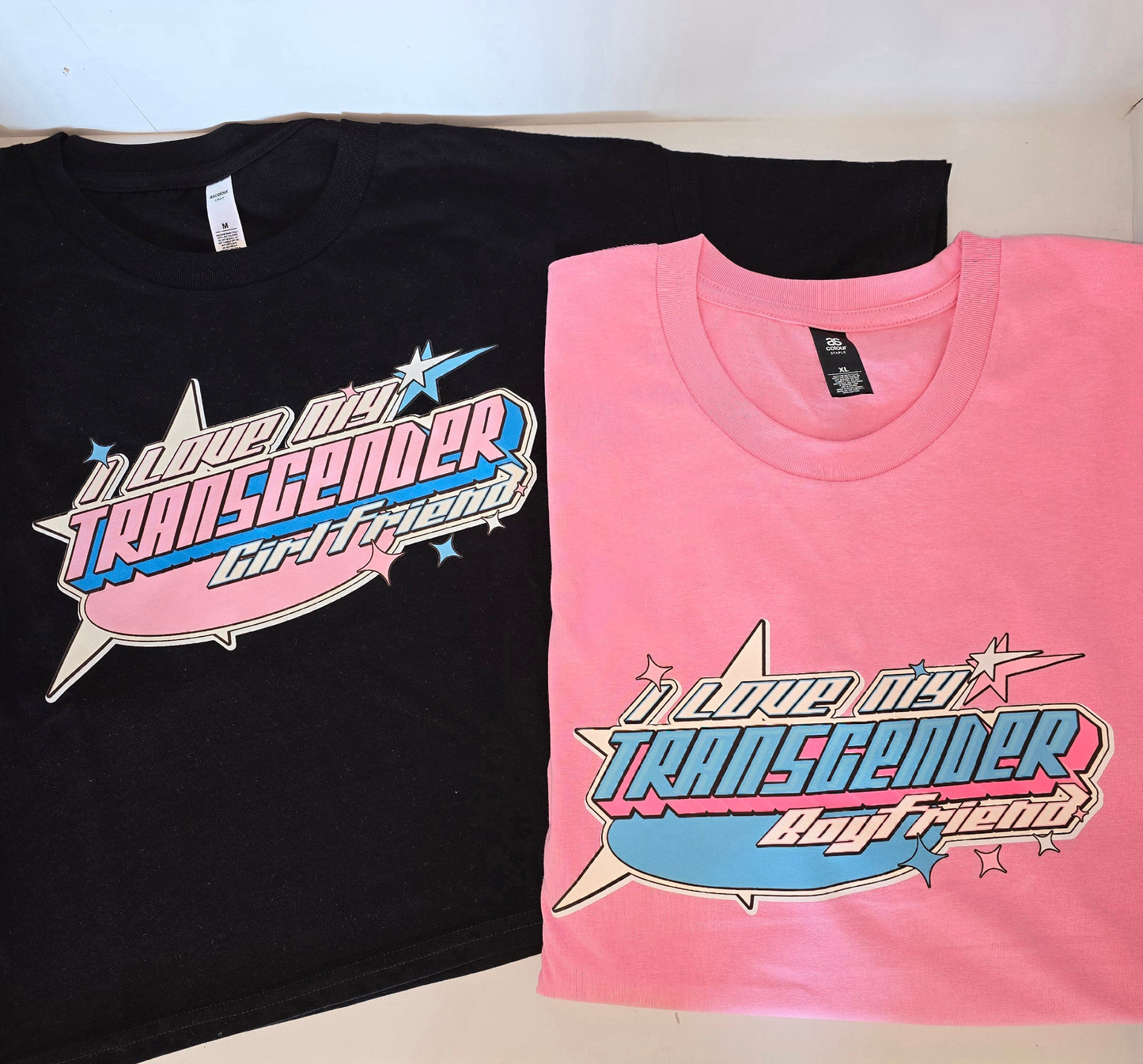 A black t-shirt designed by the small, trans and queer-owned business, Gay Skate, features a colorful front design with stylized pink and blue text reading "I LOVE MY TRANSGENDER GIRLFRIEND" plus star and lightning bolt details.