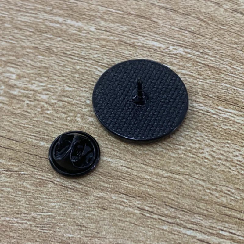 Two black plastic badges on a wooden surface: one is a textured disc with a central peg, the other is smaller with a smooth back and features the "It's A Beautiful Day To Smash The Patriarchy - rainbow and flowers" design.