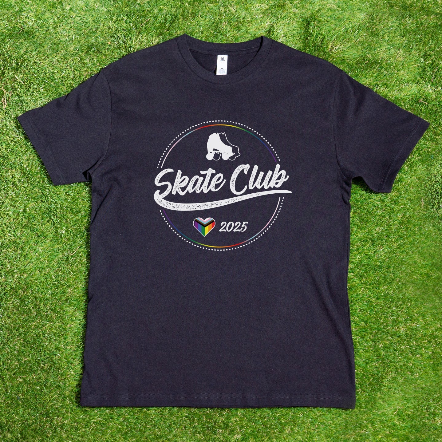 Laid flat on green grass, the Gay Skate "Pre-order: SKATE CLUB 2025 Progress Pride" T-shirt features a circular logo with a roller skate and "Skate Club 2025" text. Below, a rainbow-colored heart symbolizes inclusion, celebrating its roots as a queer and trans-owned business.