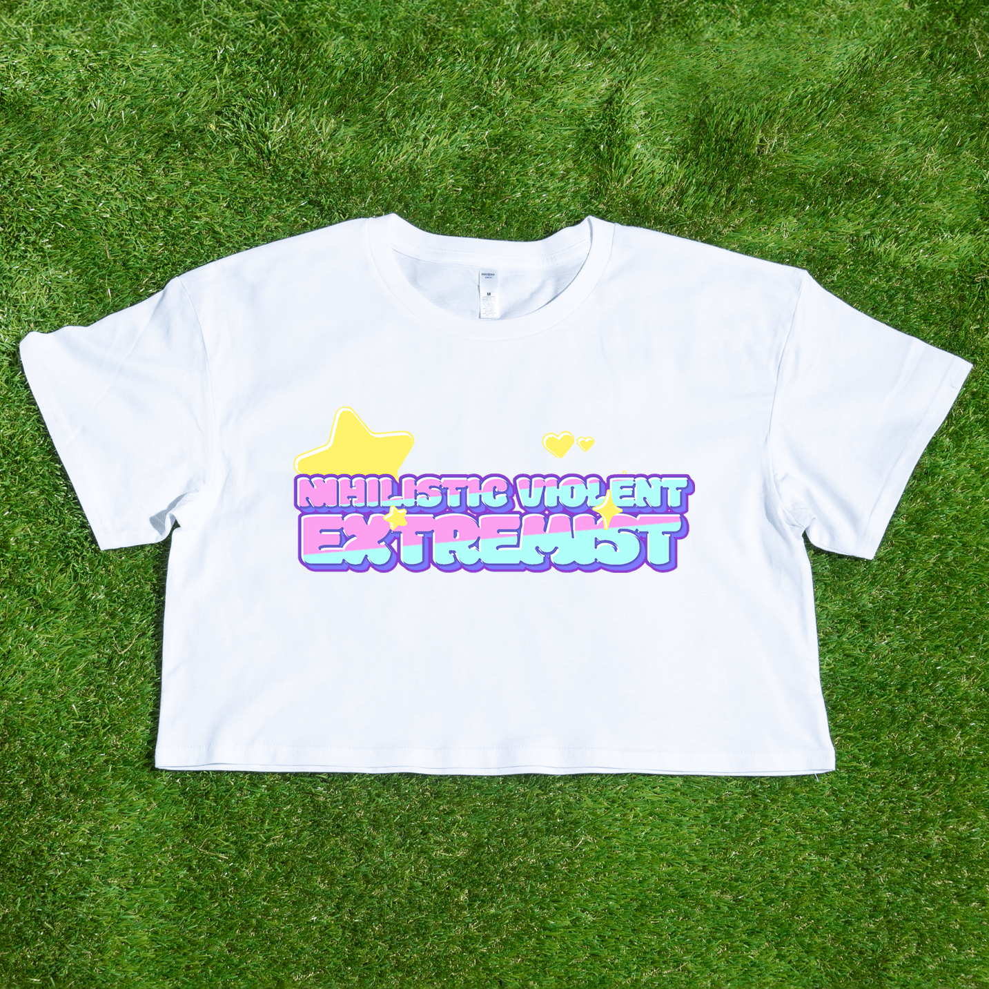The "Nihilistic Violent Extremist" locally printed satire crop tee features pastel bubble text, yellow hearts and stars on a white shirt. Hand-printed by a trans and queer owned brand and shown displayed on green grass.