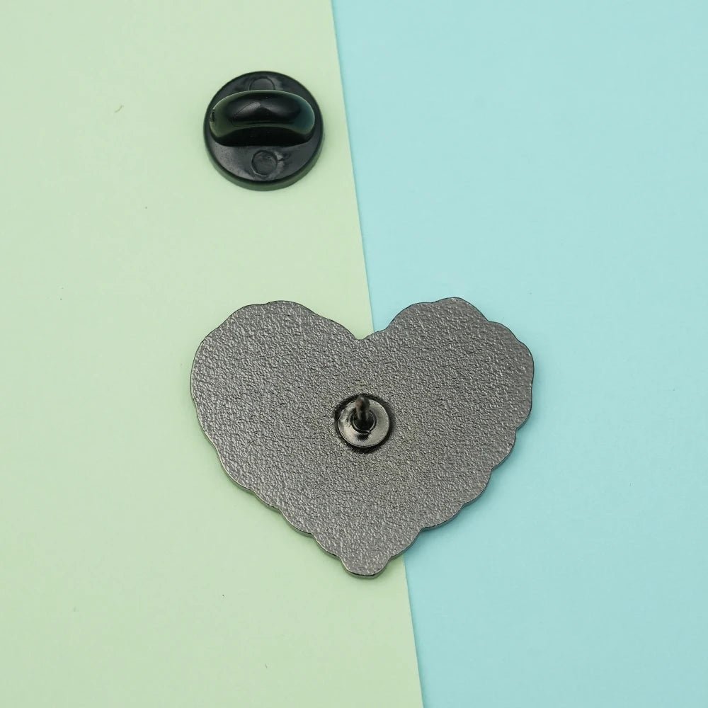The "No Human Is Illegal" heart-shaped enamel pin is shown face down on a mint and light blue background, with its black pin backing beside it—perfect for your pride accessories collection.