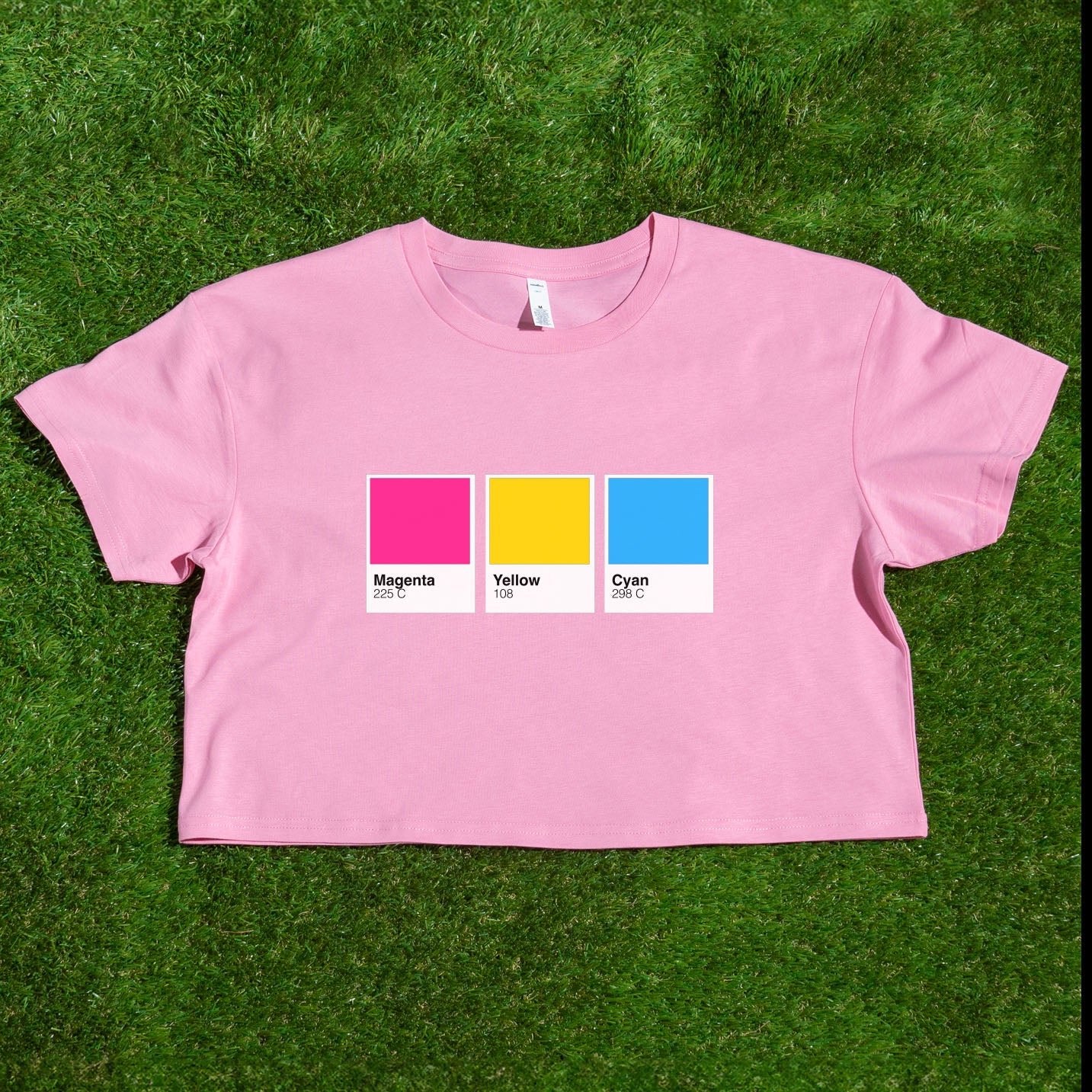 A pink crop top on grass displays Magenta, Yellow, and Cyan swatches with color codes—an eye-catching Pantone pride flag crop tee representing Rainbow, Trans, Non-binary, Lesbian, Pan, Bi, Ace & Aro; locally printed by a queer-owned business.
