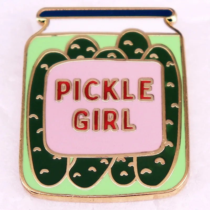 The Pickle Girl Jar Enamel Pin by Gay Skate features a vibrant pickle jar design, showcasing green pickles inside. The jar has a central label with "PICKLE GIRL" in bold red letters on a pink background. It is outlined in gold and includes a blue bar at the top to represent the jar lid—ideal for transfemmes looking to express their unique style.