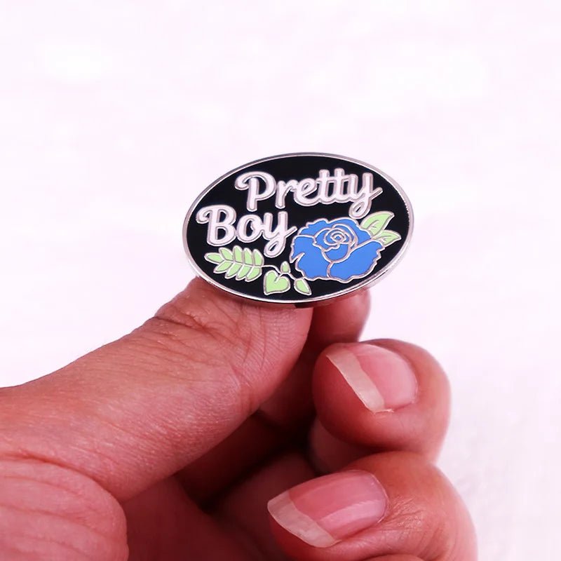 The pin, named "Pretty Boy Enamel Pin" by the brand Gay Skate, features the words in white lettering against a black background. Below the text is a blue rose detail with green leaves surrounding it. Perfect for twinks, gays, mascs, bois and other transgender and non-binary people.