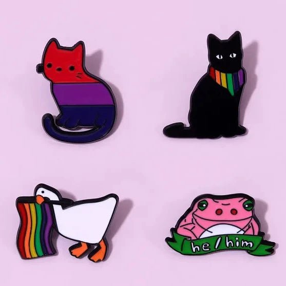 Four Pride Flag Enamel Pins from Gay Skate. A bisexual pride flag cat. Top right: a black cat wearing a rainbow bandanna. A rainbow flag waving duck. A he/him pink frog holding a "he/him" banner, celebrating queer identities.