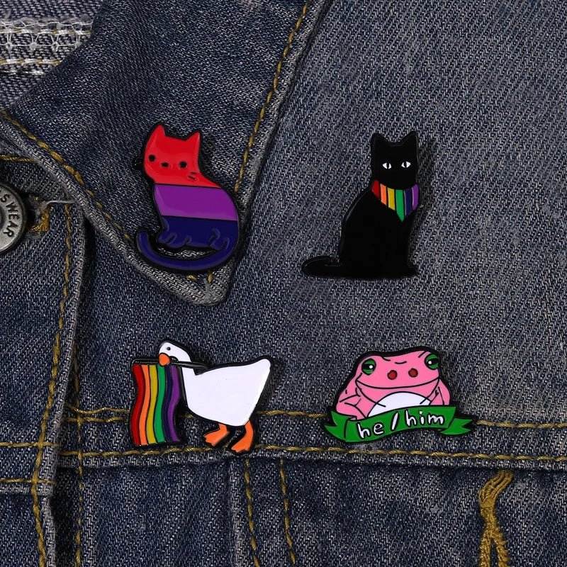 Four Pride Flag Enamel Pins from Gay Skate on a pink background. Top left: a bisexual cat. Top right: a rainbow pride black cat with a rainbow scarf. Bottom left: a rainbow duck. Bottom right: a he/him frog holding a "he/him" banner, celebrating queer identities.