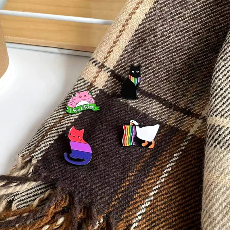 Four Pride Flag Enamel Pins from Gay Skate. A bisexual pride flag cat. Top right: a black cat wearing a rainbow bandanna. A rainbow flag waving duck. A he/him pink frog holding a "he/him" banner, celebrating queer identities.