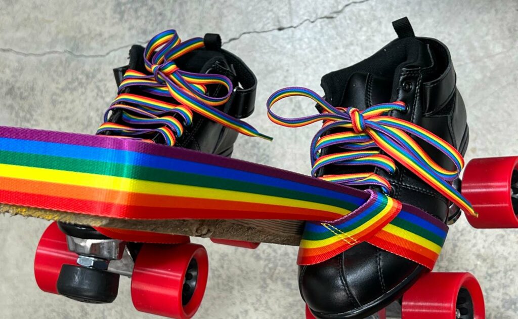 A pair of black roller skates with red wheels and rainbow-striped laces, featuring a Pride Skate Leash gear strap decorated with the rainbow flag, displayed on a concrete floor.