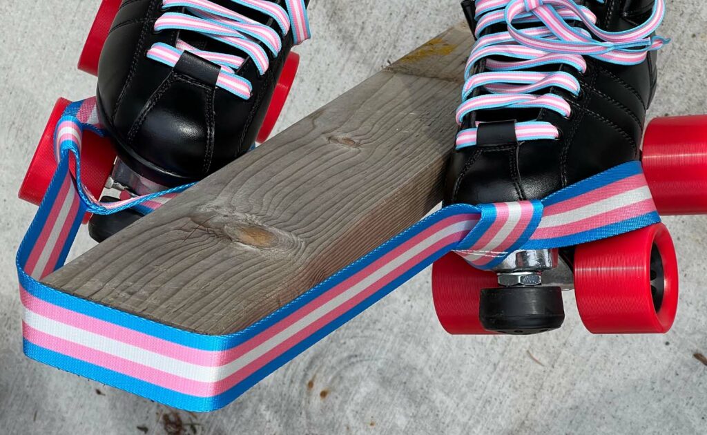 A pair of black roller skates with pink, blue, and white striped laces and matching ribbon are secured with a Pride Skate Leash – Gear strap featuring the Lesbian flag, resting on a wooden plank over concrete.