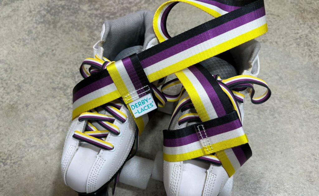 A pair of white roller skates with Pride Skate Leash gear straps in various pride flag colors rest on a gray surface. The straps, labeled "Derby Laces," include rainbow, trans, non-binary, and lesbian flag designs. Foam padding is beneath the skates.