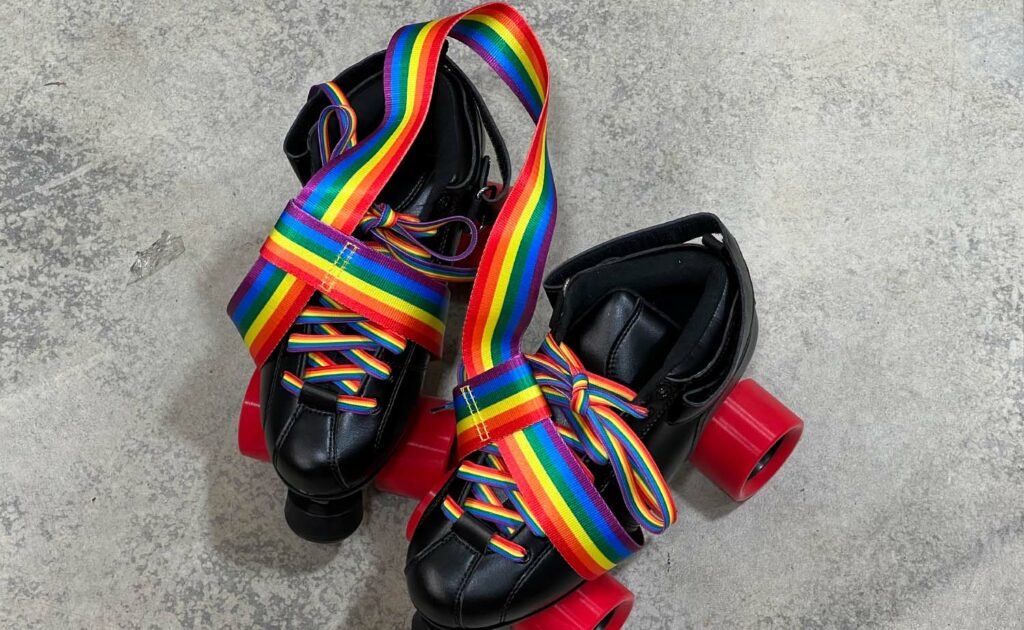 A pair of black roller skates with red wheels, rainbow laces, a rainbow-striped strap, and a Pride Skate Leash – Gear Strap featuring multiple pride flags rest on a gray textured floor.