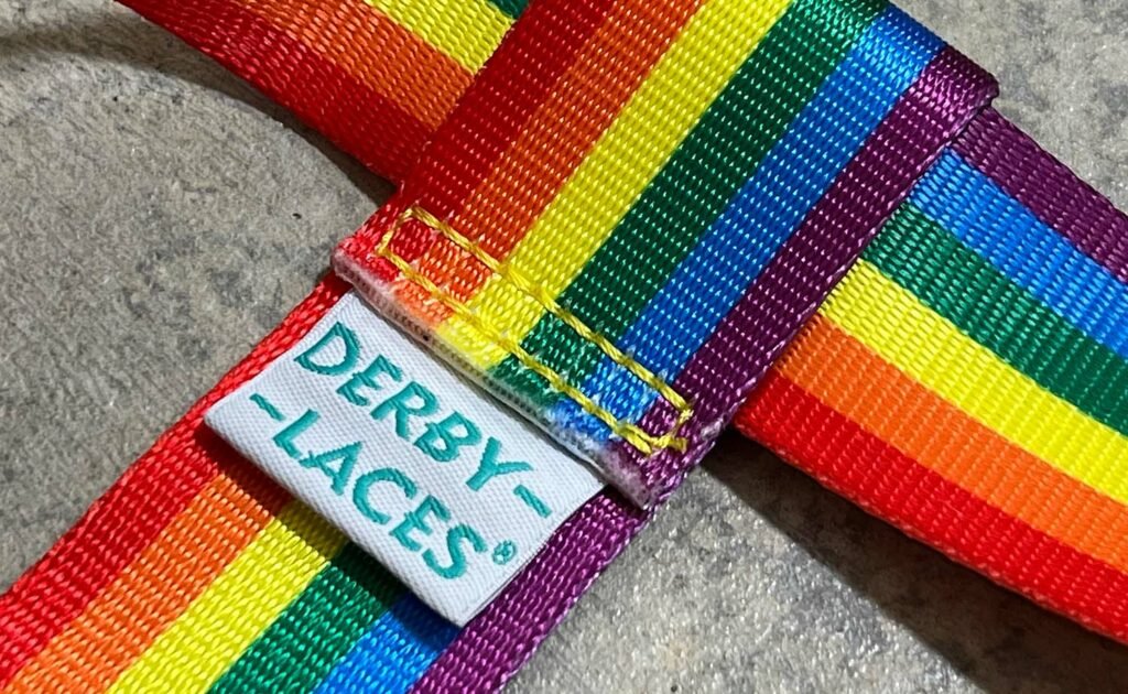 Close-up of a Pride Skate Leash gear strap featuring rainbow-striped colors inspired by the Pride flag, with a white "DERBY LACES" label in teal, resting on a gray textured surface. Available in Rainbow, Trans, Non-Binary, and Lesbian flags.