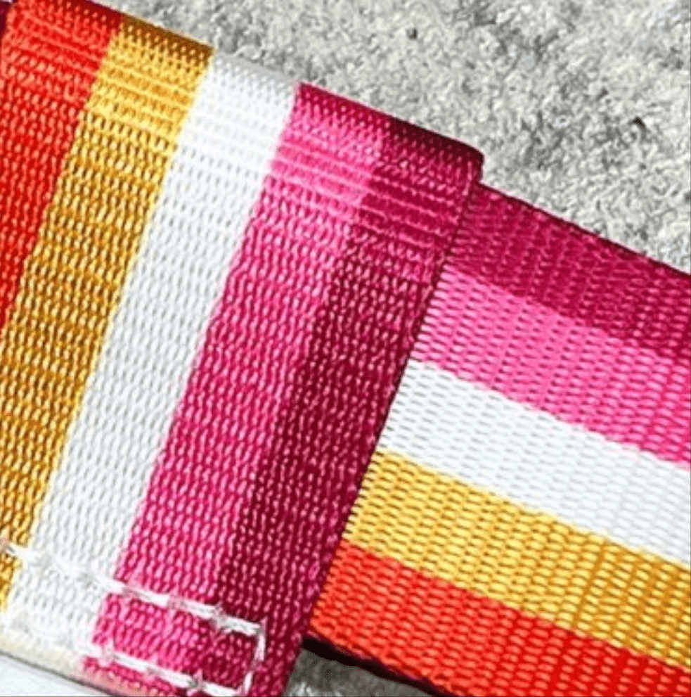 A close-up of two overlapping Pride Skate Leash gear straps, featuring horizontal stripes in red, orange, yellow, white, and pink—colors of the Lesbian flag—resting on a rough gray surface.