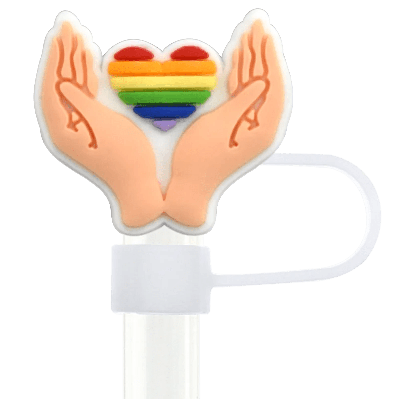 The Pride straw topper, designed to fit Stanley Cup straws, features two upward-facing hands holding a rainbow-striped heart. It's mounted on a white base with a flexible loop on the side, embodying the essence of pride designs.